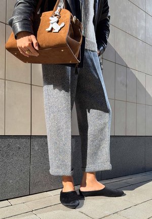 Persoană care poartă pantaloni gri cu model, pantofi slip-on negri, geacă de piele neagră, ținând o geantă maro din piele întoarsă cu breloc alb în formă de câine.