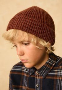 Bonnet en tricot marron avec une texture côtelée, ajusté à la base. Porté par un enfant en chemise à carreaux aux tons bleu foncé et bleu clair.