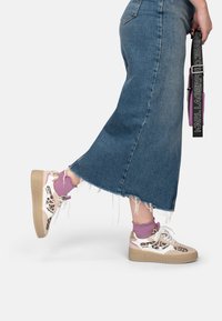 Sneakers met een beige platformzool, een bovenwerk van wit en luipaardprint, aangekleed met gouden details; gecombineerd met een spijkerrok en paarse sokken.