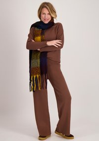 Bruine trui en broek met wijde pijpen, gecombineerd met een sjaal met textuur in meerdere kleuren, met strepen in blauw, geel en bordeauxrood. Zachte stof en een losse pasvorm.