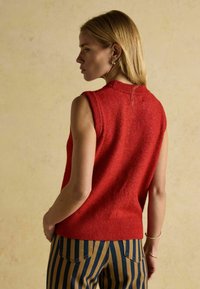 Rote ärmellose Strickjacke mit einem gerippten Kragen, aus strukturiertem Stoff. Kombiniert mit gestreiften Hosen in Beige und Marineblau. Rückansicht gezeigt.
