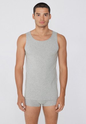 Młody mężczyzna stojący przodem, ubrany w dopasowany szary tank top i pasujące szare bokserki na jednolitym tle.