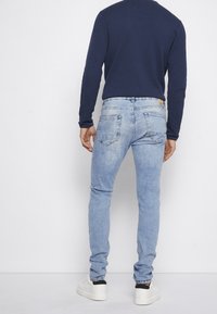 Jeans slim-fit berwarna biru muda dengan tampilan pudar dan desain lima saku. Dipadukan dengan kemeja lengan panjang navy dan sepatu sneakers putih.