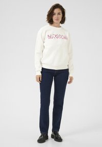 Cream sweatshirt with pink floral embroidery on the chest, paired with dark denim jeans and black shoes. Smooth texture, casual fit.