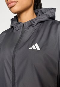 Grå hættejakke lavet af let materiale. Har en lynlås foran og hvide logoaccenter på venstre brystområde. Glat tekstur.