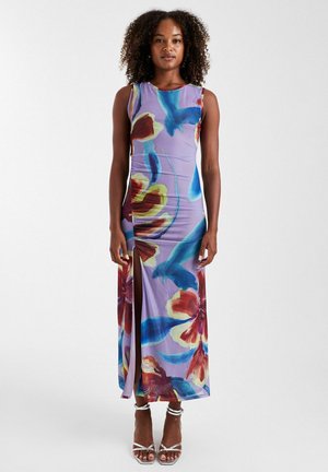 Femme aux cheveux bouclés portant une robe lavande sans manches avec de grands motifs floraux et d’oiseaux colorés et une fente latérale haute jusqu’à la cuisse.