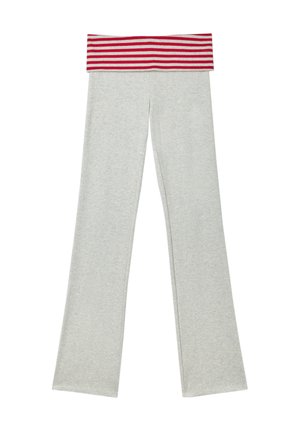 Jeune femme debout, la main sur la hanche, portant un haut à manches longues rouge, une ceinture rayée, un pantalon blanc et des chaussures claires sur fond gris.