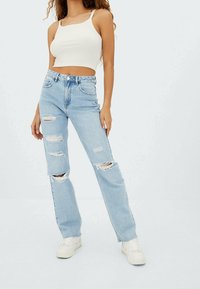 Mujer vistiendo jeans de tiro alto desgastados de color azul claro, top corto blanco y zapatillas blancas, de pie contra un fondo liso.