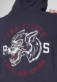 Hanorac de culoare bleumarin, cu un grafic mare reprezentând capul unui lup fioros, însoțit de litere roșii și albe care spun "PUMA STAPLE" și "O CONTAGIUNE SOCIALĂ POZITIVĂ."