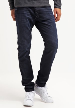 Homme portant un jean slim bleu foncé et des baskets blanches, debout avec les jambes légèrement croisées.