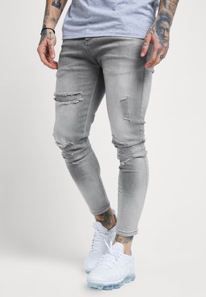 Jeans skinny de mezclilla gris con detalles desgastados, que presentan múltiples roturas y un ajuste ajustado. Combinados con zapatillas deportivas blancas.