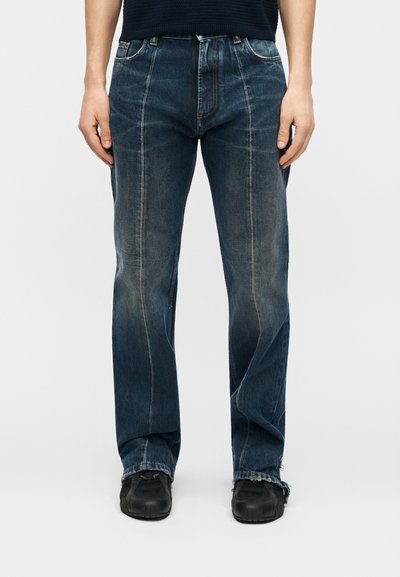 Persoană purtând blugi cu picior drept de culoare albastru închis, cu detalii de cusături frontale, și pantofi negri, stând în picioare pe fundal alb.