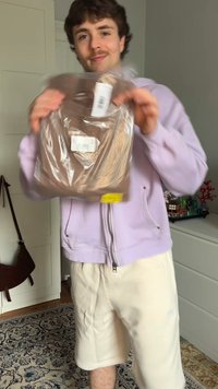 Svetlo rjav T-shirt v plastični embalaži, ki ga drži oseba v svetlo vijoličnem zadrge s kapuco in smetana kratkih hlačah, s vidnimi nalepkami.