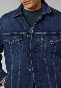 Giacca di jeans in blu scuro con un taglio classico, caratterizzata da due tasche sul petto, bottoni in metallo e dettagli di cucitura a contrasto.