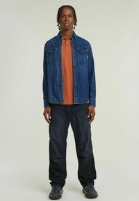 Camicia di jeans blu, polo arancione sotto, e pantaloni cargo neri; presenta tasche, chiusura con bottoni e calzature robuste.