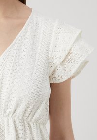 White lace top with flutter sleeves, featuring textured patterns and a cinched waist detail, showcasing intricate openwork designs.
