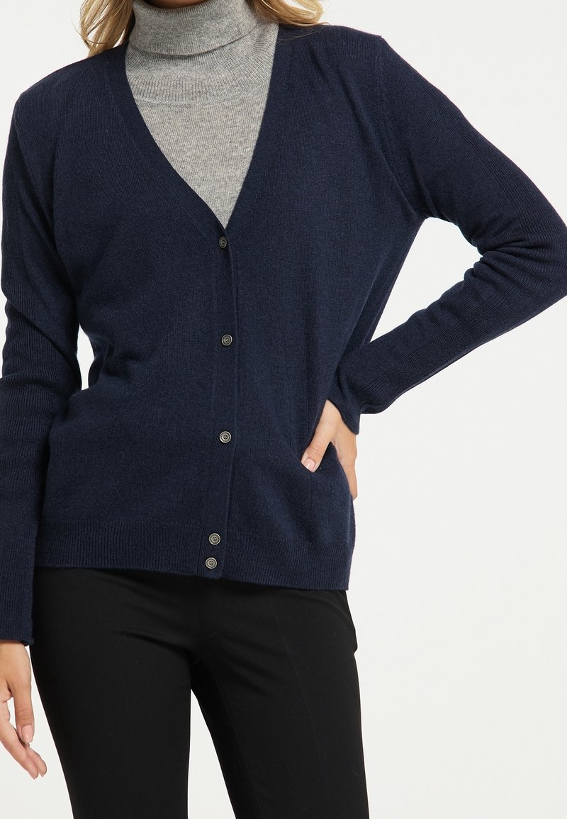 Femme portant un cardigan bleu marine boutonné sur un pull à col roulé gris et un pantalon noir, la main posée sur la hanche.