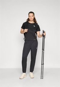 Μαύρη αθλητική μπλούζα και φόρμες από λεία ύφανση. Μαύρη σακίδιο και ραβδιά πεζοπορίας. Αθλητικά παπούτσια σε ανοιχτόχρωμο τόνο. Απλός σχεδιασμός, χωρίς σχέδια.