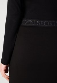 Gros plan d'une personne portant un haut noir à manches longues et une jupe noire avec une ceinture étiquetée "CAIN SPORTS."