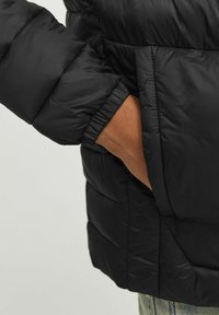 Hand in een zwarte pufferjack met elastische manchetten, vingers gedeeltelijk in de zijzak, boven lichte blauwe denim jeans.