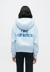 Ανοιχτό μπλε φούτερ με κουκούλα της Nike, με μεγάλο μπλε λογότυπο «nike athletics» στην πλάτη και μικρό ασημί σήμα swoosh. Διαθέτει μπροστινή τσέπη και κουκούλα.