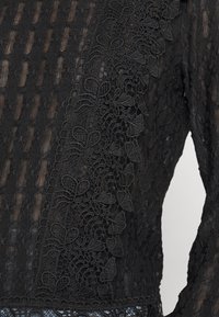 Tissu en dentelle noire avec broderie florale et bandes verticales en mesh, présentant un motif transparent et texturé sur un vêtement à manches longues.