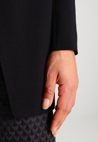 Schwarzer Strickstoffärmel mit sauberem Abschluss und leichtem Glanz, kombiniert mit strukturiertem dunkelgrauem Stoff mit Schuppenmuster.