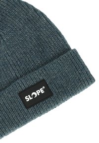 Stickad mössa i mörk teal med en vikbar ribbad kant. Har en svart rektangulär etikett med vit text som visar "SLOPE."