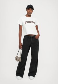 Camiseta corta blanca con el gráfico "XSURE" en marrón, combinada con jeans de mezclilla negros holgados. Accesorios incluyen un bolso gris y zapatillas blancas.