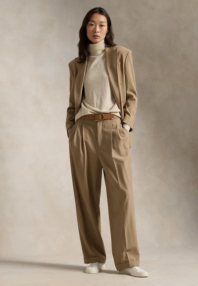 Kvinne som står med hendene i lommene iført beige blazer, matchende vidbukser, kremfarget høyhalset genser, brunt belte og hvite joggesko.