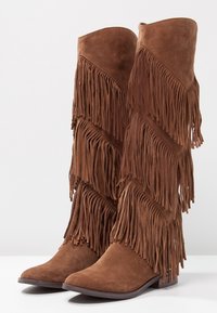 Botas altas de gamuza marrón con detalles de flecos en capas, puntera afilada y tacón plano. La textura es suave y aterciopelada.