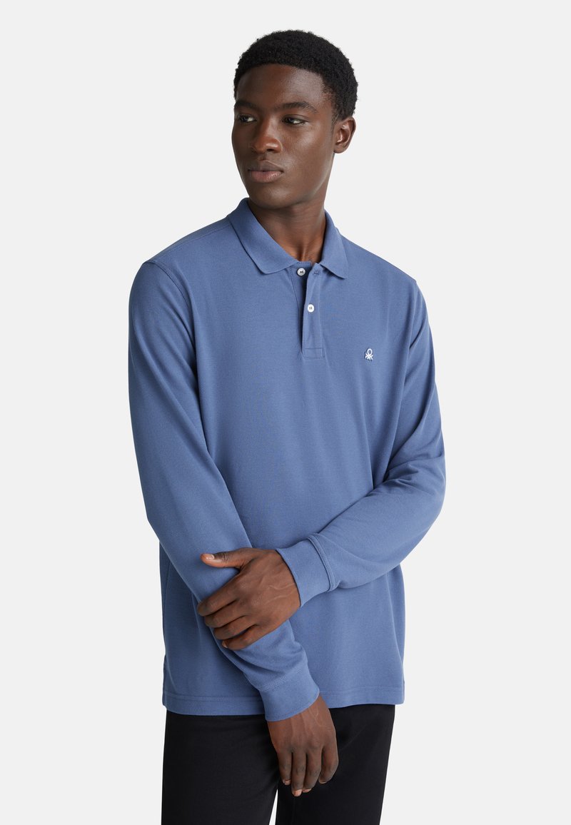 Joven con pelo rizado corto, vistiendo una camiseta polo de manga larga azul y pantalones negros, de pie con los brazos cruzados y mirando de lado.