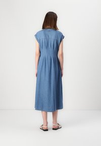 Abito di jeans con maniche corte, lunghezza fino al polpaccio, colore azzurro chiaro, dettagli plissettati sulla schiena e vestibilità comoda. Indossato con sandali piatti.