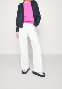 Svart lätt jacka, rosa texturerad skjorta, vita vida byxor och plattformssandaler med marinblå och metalliska accenter.