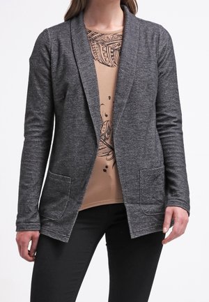 Donna che indossa un cardigan grigio scuro aperto davanti sopra una camicia beige con motivi astratti neri e pantaloni neri.