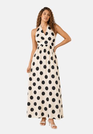 Frau trägt ein ärmelloses, Neckholder-Maxikleid in Creme mit großen schwarzen Polka-Dots und weißen Riemchenabsätzen vor einfachem Hintergrund.