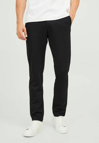 Pantaloni neri in tessuto morbido con una vestibilità slim, dotati di tasche laterali e un cinturino sottile. Abbinati a sneakers bianche.