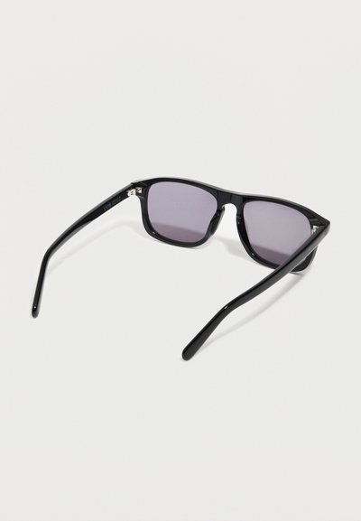 Μαύ ρα ορθογώνια γυαλιά ηλίου με σκούρους φακούς, που ακουμπούν σε λευκή επιφάνεια, με τα μπράτσα ανοιχτά προς τα έξω.