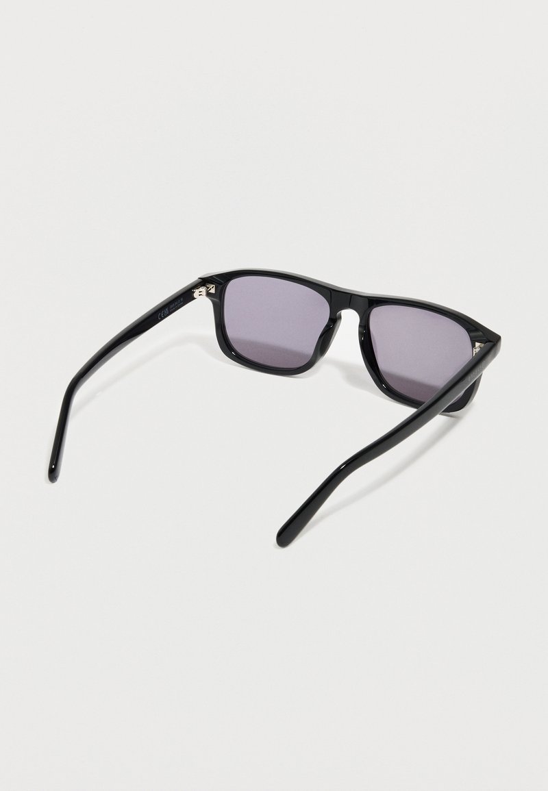 Schwarze rechteckige Sonnenbrille mit dunkel getönten Gläsern, die auf einer weißen Oberfläche liegt, die Bügel sind nach außen ausgestreckt.