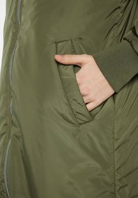 Olivgrüne, gepolsterte Jacke mit einem Reißverschluss. Verfügt über eine Seitentasche mit genähtem Rand. Hat einen gerippten Ärmelbund für zusätzliche Details.