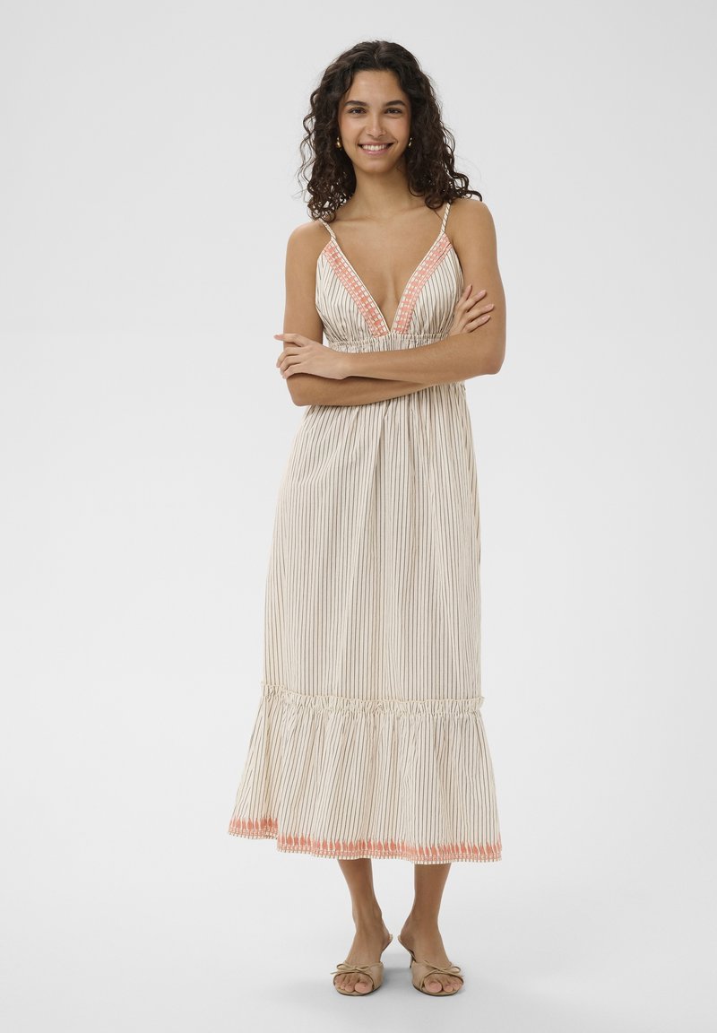 Femme aux cheveux bouclés portant une robe maxi sans manches rayée avec une broderie orange, debout pieds nus, les bras croisés et souriante.