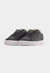 Paire de baskets basses noires avec semelles blanches, lacets noirs, œillets dorés et logo Tommy Hilfiger sur le côté.