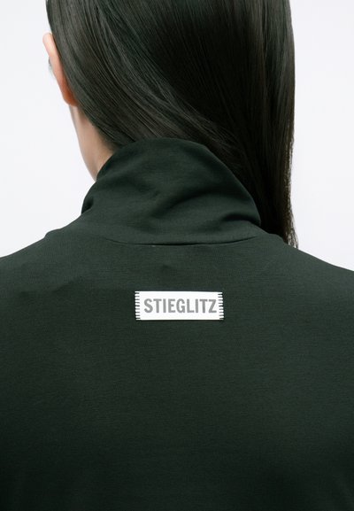 Tmavozelená, priliehavá mikina s vysokým golierom a obdĺžnikovou bielou etiketou s nápisom "STIEGLITZ" na chrbte. Hladká textúra látky.