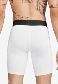 Pantaloni scurți albi de sport, cu un brâu elastic negru, având buzunare laterale și un material neted și elastic. Vederea din spate arată ajustarea și lungimea.