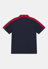 Polo tričko s krátkým rukávem v námořnické modři, s červenými rameny a černým textem po stranách, zobrazeno zezadu na bílém pozadí.