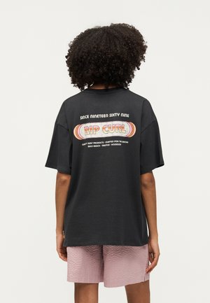 Άτομο με σγουρά μαλλιά στέκεται με την πλάτη, φοράει μαύρο μπλουζάκι Rip Curl και υφασμένα ανοιχτό ροζ σορτς.