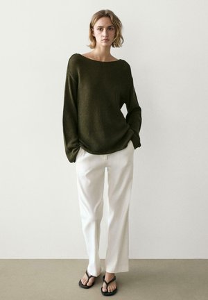 Femme portant un pull en maille oversize vert foncé, un pantalon blanc à jambes larges et des sandales noires, debout devant un fond clair uni.
