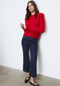 Rdeča puhasta pletena jopica z dolgimi rokavi, v kombinaciji s temno modrimi razširjenimi džinsom z kontrastnim šivanjem in črnimi balerinkami. Minimalistična ozadje.