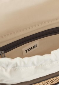 Interior de bolsa de lona beige con un bolsillo con cremallera oscura. Presenta un borde de tela blanca fruncida y una pequeña etiqueta "TOUS" en el costado.