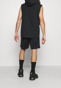 Black sleeveless hoodie with a hood, paired with black shorts. Worn with black textured socks and sneakers featuring a speckled sole.